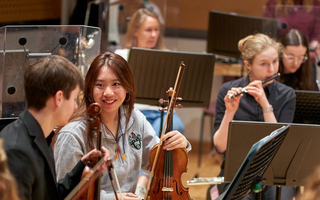 Two violin players are talking to each other. Behind them is a piccolo flautist.