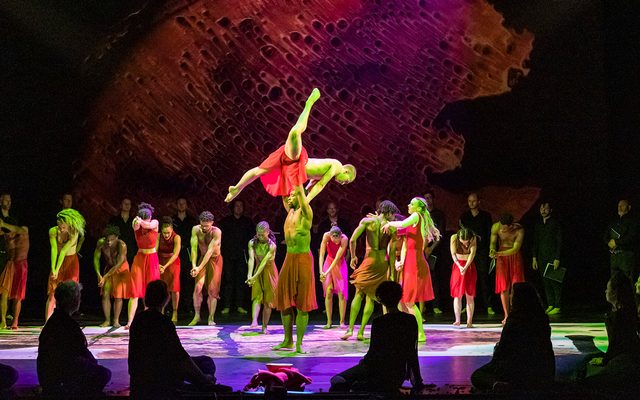 A group of dancers on the stage wearing reds and oranges. in the centre a man is holding another man above him.