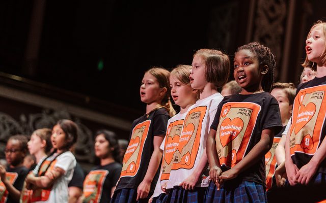 A large group of children wearing matching t-shirts featuring a playing card motif, in an ornate theatre