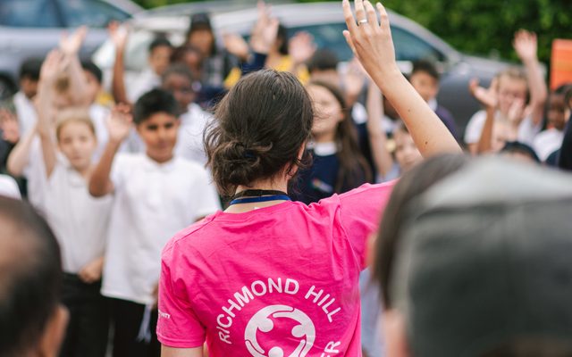 A lady in a pink t-shirt with the wording Richmond Hill in white on the back stands facing a group of children with her right hand in the air