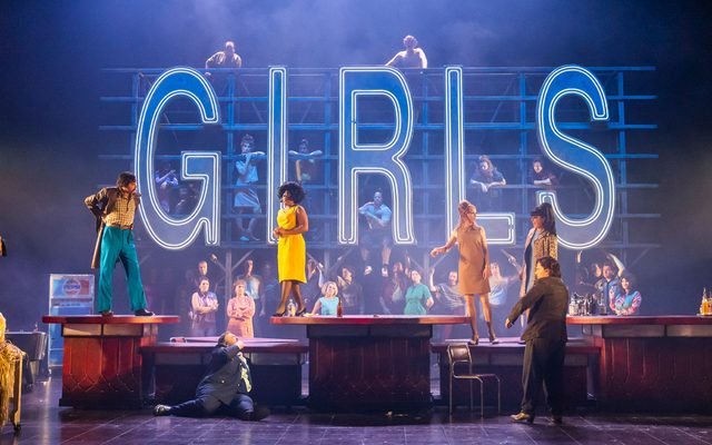 Patrons of a club walk the stage, a neon sign of the word 'GIRLS' can be seen in the background.