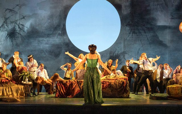 A woman in a green gown is stood facing away with her arms outstretched. In front of her a party is happening.