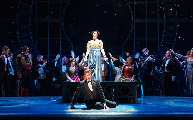 A man is on the floor in the middle of the stage. Behind him is a woman in a blue gown there are two men in suits either side of him. Behind them is a large crowd.