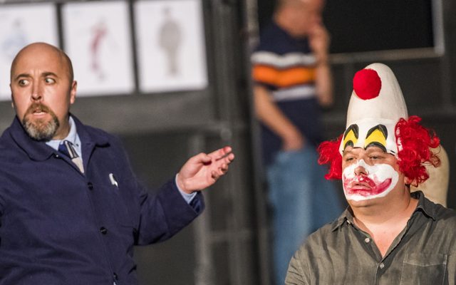 A man with clown make up is sat on a chair. Beside him is a man in a suit.