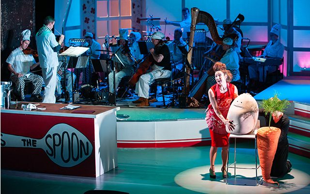 A woman wearing a red dress, standing on stage next to a giant egg and carrot in the Pass The Spoon show