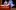 Person dressed as Pinocchio with long wooden nose sits on a bed and looks at a blue fairy who holds up a finger.