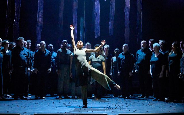 A woman dancing, she is on one leg with her arm raised up. She is surrounded by singers.