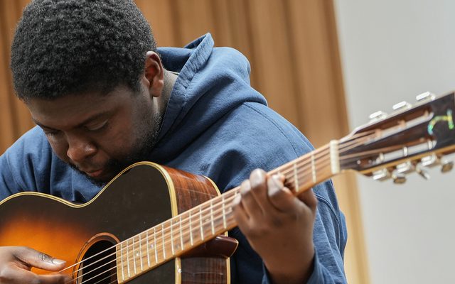 A man plays an acoustic guitar in a wood-panelled studio with a microphone to his left.