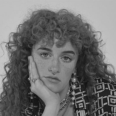 A woman with long curly hair, a nose ring and an eye brow piercing.