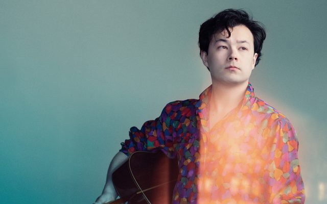 A young man in a colourful shirt holds a classical guitar under his arm, and looks off to his left, against a light blue background