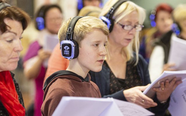 A young boy wears feedback headphones and holding sheet music, singing in a community choir