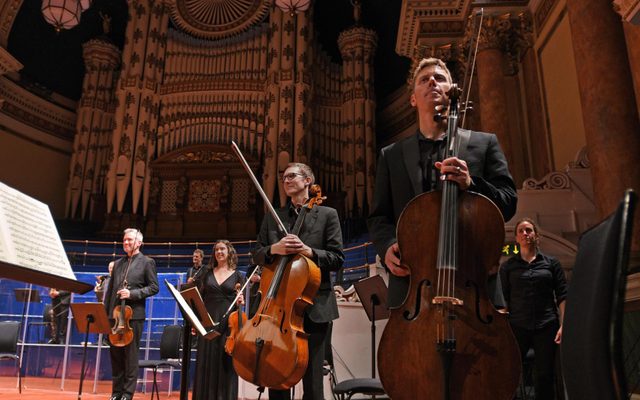 An orchestra stands with instruments in a grand concert hall setting at Leeds Town Hall
