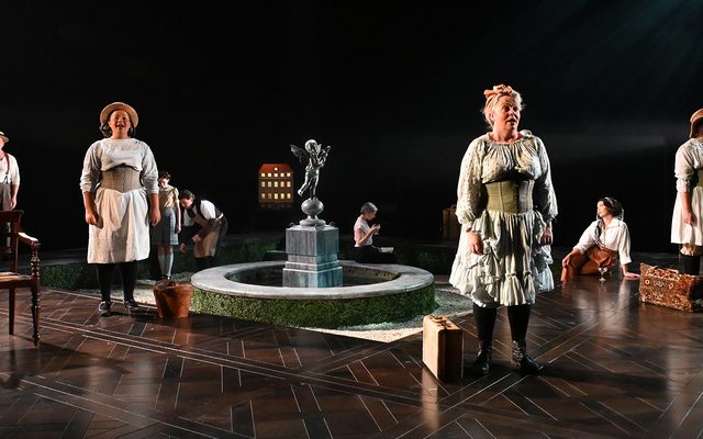 Three women and a man dresses in 18th century style under-garments stand equally distanced on a stage, singing. A number of props including a chair and a fountain can be seen behind them.