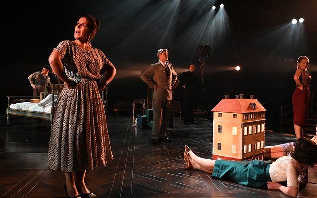 Two young women recline on the floor looking at a dolls house while others stand around them singing