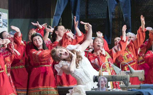 A group of people dressed in red behind a woman in a white dress. She is at a sewing machine.