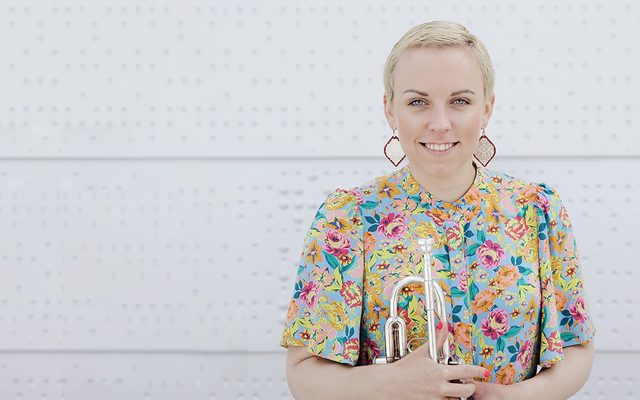 A smiling woman in a bright floral dress stands against a patterned white wall