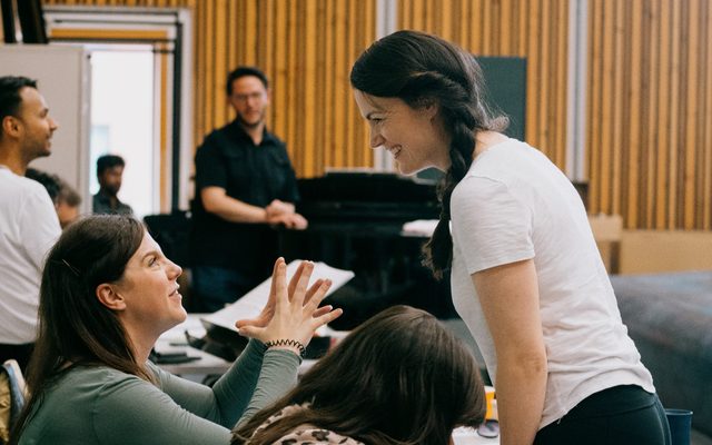 Two woman are looking at each other smiling, there are a few people and a piano behind them