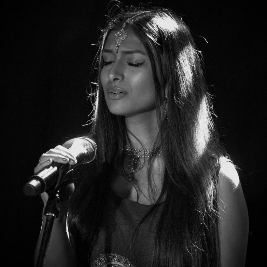 Ashnaa Sasikaran A woman singing into a microphone. She has long dark hair and is wearing jewellery around her neck and on her forehead.