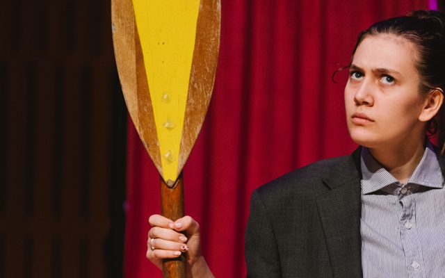 A white teenage wearing a lawyer's gown stands holding a yellow oar looking annoyed