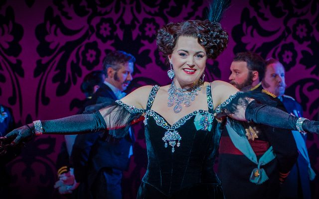 A dark-haired woman in a glamorous velvet dress with jewelled trim smiles at the camera and stretches out her arms