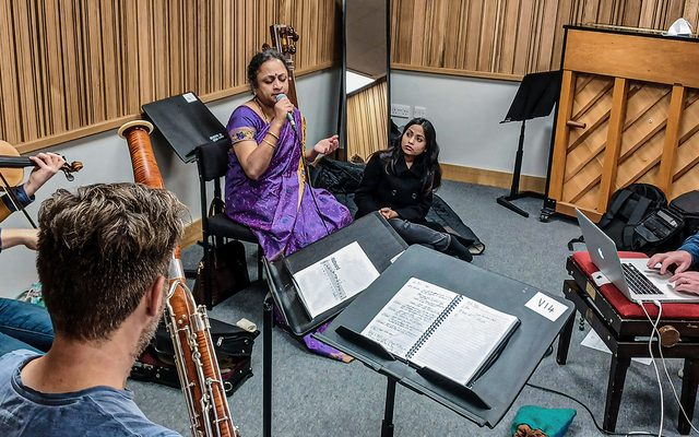 A woman in a purple sari sitting in the centre of a group of musicians rehearsing in an acoustic wood-panelled studio