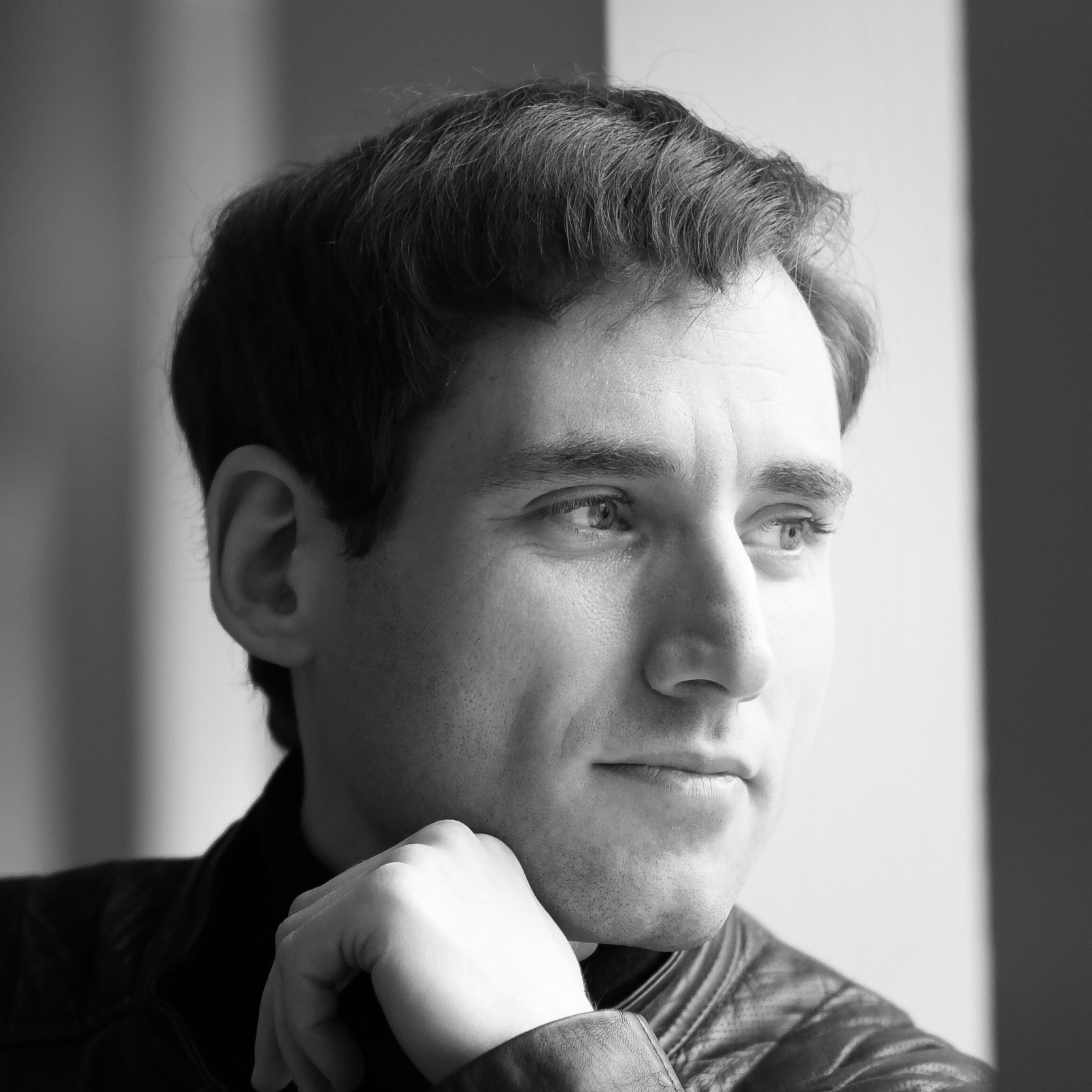 A man with short dark hair looking to the right. One of his hands is raised to his face.