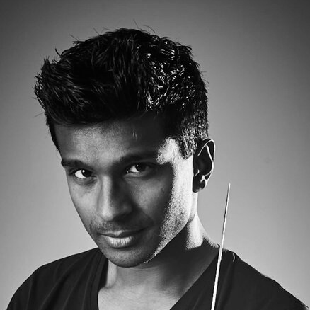Leslie-Suganandarajah A man with short dark hair wearing a t-shirt and holding a conductor's baton.