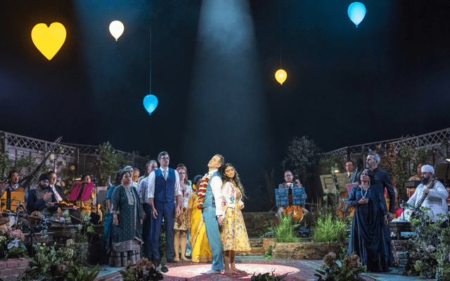 A man in a blue suit and a woman in a floral dress are stood in the middle of the stage with a spotlight on them. They are surrounded by people and there are lit up balloons in the sky.