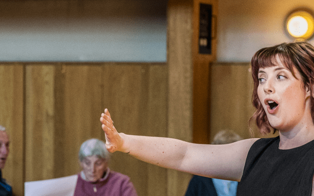 A woman holding out her hand whole singing. Behind her there are some people sat.