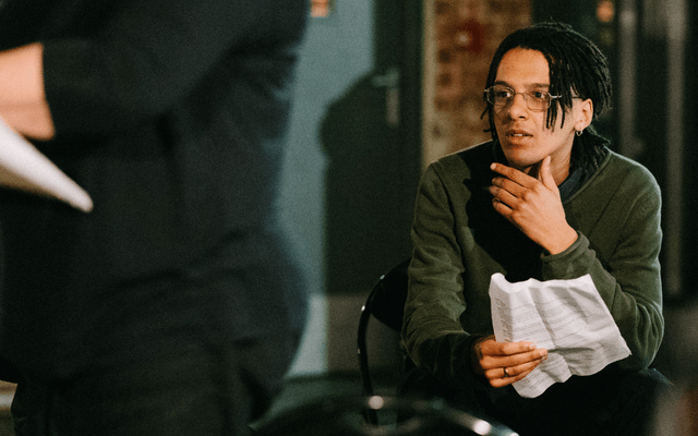 A man with black hair, silver glasses and a green top, holds a sheet of paper whilst taking part in a music writing workshop at Opera North.